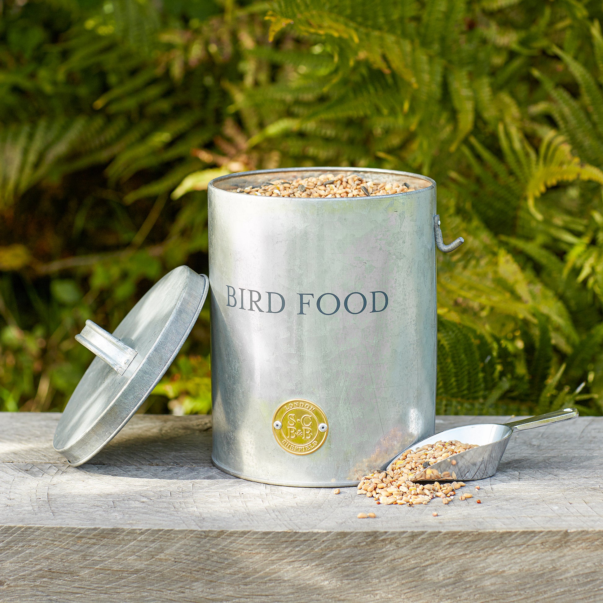 Galvanised steel bird food tin full of seeds, with its lid off and scoop next to it and placed on a wooden bench with green foliage in the background.